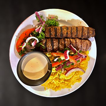 Two grilled beef kebab skewers over yellow rice with salad and tahini